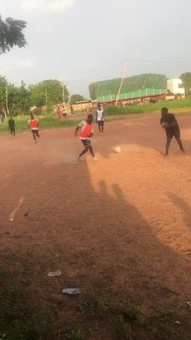 This media file depicts a group of young people playing soccer on a dirt field in Wukari, Nigeria. The subjects are primarily young men, many of whom are wearing athletic clothing, including shorts and t-shirts or training bibs in red and white. One individual is wearing a black t-shirt and shorts, while another is in a pink top and black shorts. A white soccer ball is visible in play.

The scene is an outdoor, rural or semi-rural setting. The ground is a reddish-brown dirt, with patches of dry grass and some scattered debris in the foreground, including plastic bottles and bags. In the background, there are trees, some buildings, and a large truck with a green tarp covering its load. The sky is visible, suggesting it is daytime, likely late afternoon given the long shadows cast across the field. The overall mood appears to be one of casual play and recreation. There is no visible text in the media file.