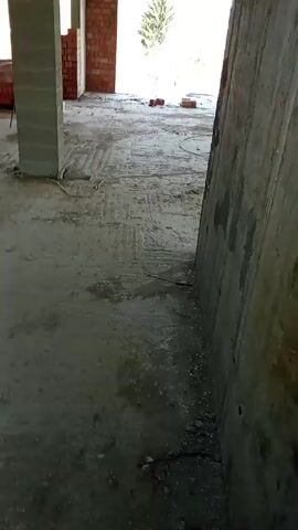The video shows the inside of a building under construction. The space is largely bare, with a concrete floor and unfinished concrete pillars and walls. There are a few bricks and some electrical wiring scattered around. Looking through the windows, we see a construction site with wooden planks, metal pipes, and construction debris. A person is visible in the distance, working.