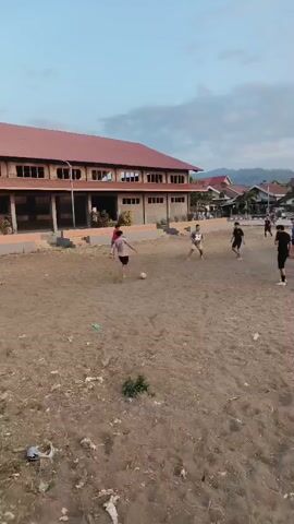 The video shows an aerial view of a group of young people playing soccer on a dirt field. The field is rough and uneven, with some debris scattered on the ground. In the background, there is a large building with a red roof, as well as several smaller houses and some trees. The sky is cloudy, and the lighting suggests it is either early morning or late afternoon. There are at least seven people visible on the field, all dressed casually. They are actively engaged in the game, kicking and running after a soccer ball. The camera angle is high and slightly tilted, giving a wide perspective of the activity.