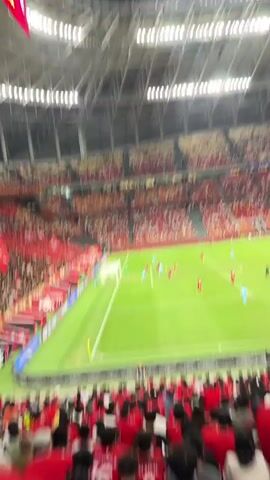 The video shows an overhead view of a soccer field in a stadium during a soccer match. The field is green and well-maintained. One team wears red uniforms, and the other wears light blue. Players are actively engaged in gameplay near the goal. The goalie, dressed in green, is in position.  Spectators fill the stands, predominantly wearing either red or light brown. The upper tiers of the stadium are visible, indicating a large seating capacity. The camera is positioned high above the field, likely in the stands, providing a wide view of the action.