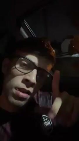 The video shows a young man with short brown hair, wearing black-rimmed glasses and a dark-colored shirt with a logo, sitting in a car at night. He is looking at the camera and gesturing with his fingers. Music is playing in the background. The interior of the car is dark, and through the window, you can see streetlights and possibly a building in the background.