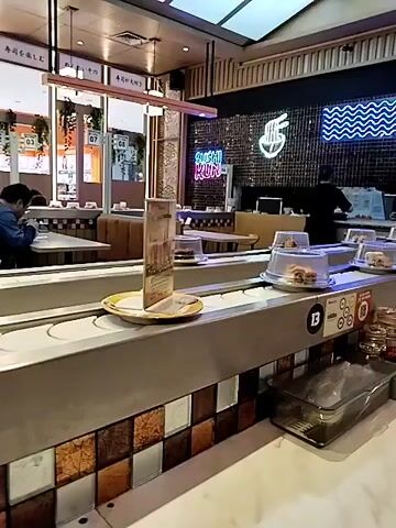 The video shows a conveyor belt sushi restaurant. The belt is moving, and there are several plates of food on it, each covered with a clear plastic lid. Some of the plates appear to contain burgers or sandwiches. There are also customers seated at tables in the background. A sign on the wall says "SUSHI HUT." The restaurant is well-lit and appears to be clean and modern.