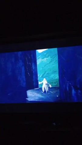 The video displays a scene from a film, likely a fantasy movie, on a projector screen. The setting appears to be a dramatic, otherworldly landscape with large, dark, vertical structures framing the view. In the foreground, a figure wearing a long, flowing white robe and with long white hair is prominent. This individual holds a staff that emits light at the tip. The figure is in motion, seemingly gesturing or casting a spell, with their arms outstretched and their robe billowing. The background shows a vista of mountains and a cloudy, blue sky. The lighting is dramatic, with the primary light source appearing to come from the staff and the sky. The overall mood is epic and magical.