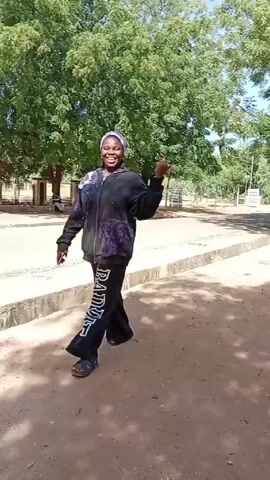 The video shows two young women outside. The first woman is standing in the shade of some trees. She is wearing a purple headscarf, a black and purple hoodie, and black pants with white writing down the side. She raises her hand and makes a gesture, then turns and walks away. The second woman is walking down the sidewalk. She is wearing a black headscarf, a white, black, and green jacket, and maroon pants. She is also carrying a cell phone. The environment is a park-like setting with trees, a sidewalk, and a fence.
