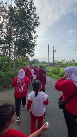 A group of people, mostly wearing red shirts and some with headscarves, are walking down a paved road. Some of the individuals are children, also dressed in red. The road is lined with lush green vegetation, including trees and what appears to be a rice paddy. The sky is partly cloudy. In the background, a person is riding a motorcycle. The camera is positioned behind the group, following them as they walk away. One of the children in the foreground is holding a white object, possibly an egg, and extending their arm. The overall atmosphere appears to be a casual outdoor activity or procession.