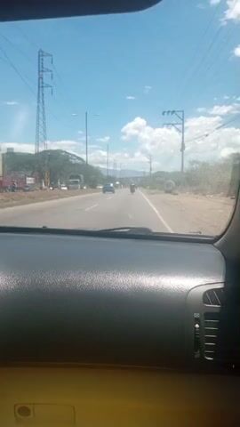 The video shows a view from inside a car, looking out through the windshield. It is a bright, sunny day with a clear blue sky and some scattered white clouds. The car is on a paved road, likely a highway or a main road, with multiple lanes.

In the distance, there are other vehicles on the road, including cars and motorcycles. The road is flanked by vegetation, trees, and bushes on both sides. In the background, there are hills or mountains. Power lines and utility poles are visible overhead and along the side of the road.

On the left side of the road, there appears to be a large truck parked. On the right side, near the edge of the road, there is a large, round, white object that looks like a spare tire or a similar industrial item.

The camera is positioned behind the dashboard of the car, which is dark gray and textured. The view is slightly tilted, suggesting the car is in motion. The overall impression is a typical daytime drive on a road in a semi-rural or suburban area with infrastructure for electricity transmission.