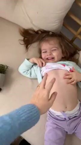 A young girl, appearing to be a toddler, is lying on her back on a light-colored surface, possibly a bed or couch. She is wearing a light blue long-sleeved shirt with cartoon designs, lavender pants, and a small pink hair clip. An adult hand, wearing a blue knitted sweater and a gold ring, is tickling the girl's stomach. The girl is laughing hysterically with her mouth wide open, exposing her teeth. Her brown hair is spread out on the surface beneath her. The background includes a white pillow and a wooden structure, potentially part of a crib or bed frame.