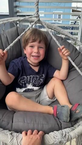 A young boy with brown hair and blue eyes is sitting in a gray, netted hanging chair. He is wearing a dark blue t-shirt with white lettering that reads "SURF" and gray shorts. He has a dark beaded necklace around his neck and is wearing dark gray Croc-like shoes with pink accents. The boy is smiling widely and holding onto the ropes of the chair with both hands, looking towards the camera.

The scene appears to be outdoors on a porch or deck, with a slatted wooden fence and a building with blue siding visible in the background. The lighting suggests it is daytime, likely with mild weather, as the boy is dressed for comfort outdoors. A person's hands are visible at the bottom of the frame, possibly pushing the chair. The hanging chair is padded with a gray cushion. The overall mood is playful and happy, with the boy appearing excited. The video clip shows the chair gently swinging, with the boy looking engaged and delighted.