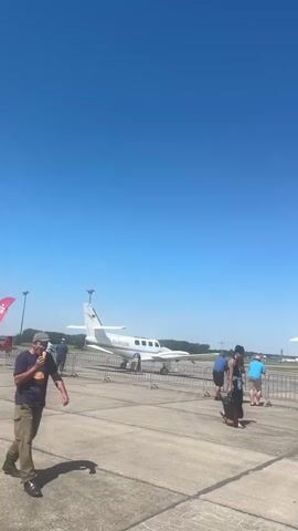 The video opens on a sunny day with a clear blue sky. The camera is positioned on an asphalt surface, likely an airfield or tarmac. In the foreground on the left, a person in a t-shirt and camouflage pants is standing, looking to their left and speaking. Further into the scene, several small airplanes are parked on the tarmac. A white twin-engine propeller plane with the registration D-ISCA is visible on the left. People, including adults and children, are gathered around this aircraft, some appearing to be observing or interacting with it. A temporary fence is set up around the aircraft. In the mid-ground and background, more people are seen walking across the tarmac. Several tall light poles are visible across the airfield. A red banner with white text is partially visible on the right side of the frame, suggesting an event or exhibition. Another white airplane is partially visible on the far right. The overall impression is an outdoor event or open day at an airfield, with spectators interacting with parked aircraft.