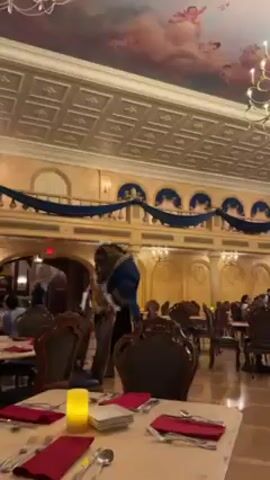The video shows a character dressed as the Beast from Beauty and the Beast standing in a dining room. The Beast is wearing a blue coat with gold trim and has a furry head and hands. He is holding something in his hands. The dining room has tables set with silverware and red napkins. There are other people in the background, including a man in a plaid shirt walking behind the Beast. The room is decorated with blue and gold trim, and the ceiling has a mural of angels.