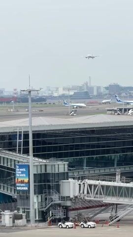 The video shows an airport scene. In the foreground, there is a modern airport terminal building with large glass windows and a jet bridge. Several vehicles, possibly luggage tugs, are parked on the tarmac near the terminal. In the middle ground, several commercial airplanes are visible on the runway and taxiways. One plane is a Boeing 787 with ANA livery. In the background, a cityscape with tall buildings is visible under a cloudy sky. An airplane is seen in flight in the upper portion of the frame, descending towards the airport. The overall atmosphere appears to be that of a busy airport during the daytime.