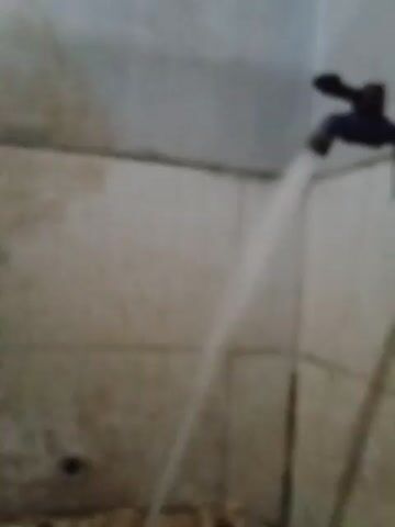 The video shows a stream of water spraying out of a blue faucet fixture toward the floor. The floor is tiled with large, square, light gray tiles. The water stream hits an area to the right, near a partially visible structure with a mottled brown and white surface. A small, round drain is visible in the floor near the right edge of the frame. The wall on the left side of the frame is a plain, light gray. The scene is brightly lit.