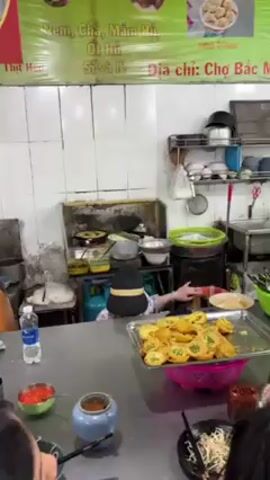 The video shows the interior of a bustling food stall in Vietnam. A person wearing a black hat and striped shirt stands behind a counter, preparing food. On the counter, there's a large tray filled with what appears to be fried egg cakes, along with various bowls and containers with ingredients and sauces. Customers are seen in the foreground, eating from bowls. The kitchen area is visible behind the counter, with various cooking equipment, utensils, and supplies. The stall's signage, "Quan TAM," is displayed on the wall, along with a list of dishes and a phone number. The overall atmosphere is lively and suggests a busy food vendor serving customers.