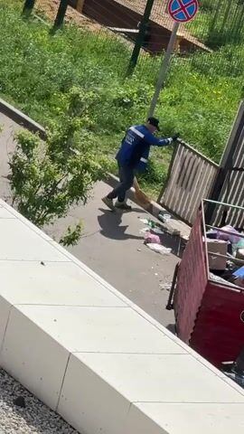 The video shows a man in a blue work jacket, jeans, and a black cap sweeping debris into a red dump truck. The truck is parked next to a fenced area with overgrown vegetation. A traffic sign indicating "no stopping" is visible in the background. The man is using a broom and appears to be cleaning up litter. The scene is shot from an elevated perspective, possibly from a building.