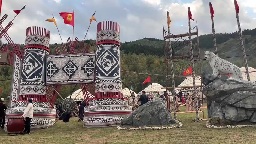 The video shows a festive outdoor scene with traditional yurts in the background. In the foreground, there is a large, ornate gateway decorated with geometric patterns and flags. A statue of a snow leopard on a rock sits to the right of the gateway. Several people are visible, some dressed in traditional clothing. One person on the left is playing a large drum. The overall atmosphere appears to be that of a cultural event or festival.
