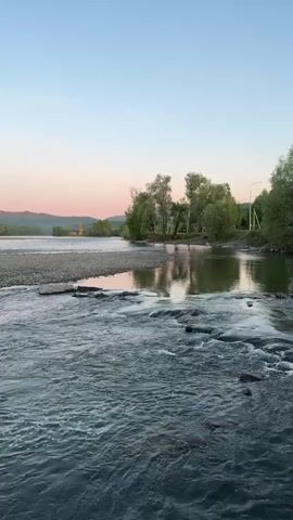The video shows a wide river flowing rapidly with small rapids over rocks. The riverbanks are lined with trees, and a gravel bar extends into the river. In the distance, there are hills and a faint golden building is visible. The sky is a clear gradient of blue to pink, indicating either sunrise or sunset. There are no people visible in the video.