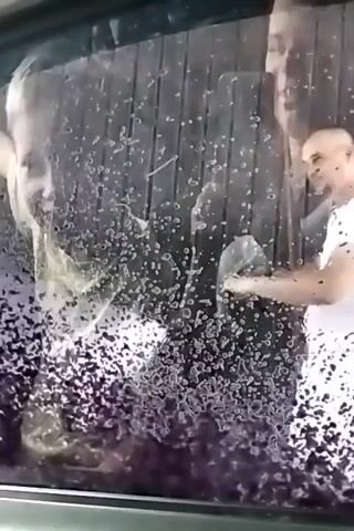 The video shows a view through a car window that is covered in purple foam. The foam is distributed unevenly across the glass, with some areas completely obscured and others showing through. Through the foam, people can be seen inside the car. At the beginning of the video, a person's head is visible, possibly looking towards the camera. Later, another person appears to be holding a phone and taking a photo or video. A young boy with blonde hair is then seen reaching out to the window and touching it. His hand is partially visible as it makes contact with the glass. Finally, another person, wearing a patterned garment, is seen inside the car, with their hand on the interior door panel. The overall impression is of a car undergoing a wash, with the interior occupants visible behind the foamy exterior.