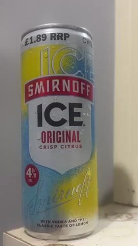 A close-up, eye-level shot captures a chilled can of Smirnoff Ice, Original Crisp Citrus flavor. The can is silver, yellow, and light blue, with numerous water droplets visible on its surface, indicating it has been refrigerated. The prominent branding "SMIRNOFF ICE" is displayed in large black letters, with "ORIGINAL CRISP CITRUS" written below it in red text. A red circle on the lower left of the can indicates "4% VOL." At the top, "£1.89 RRP" is visible, along with a partially visible "CHILL" instruction. The bottom of the can features "Smirnoff" in a script font and text reading "WITH VODKA AND THE CLASSIC TASTE OF LEMON." The background is a plain, light-colored wall. No people are visible, and the can appears to be resting on a white or light-colored surface.