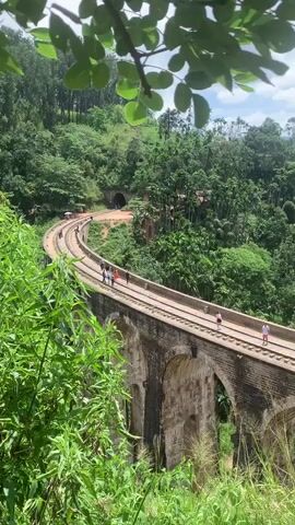 A vertical, long shot captures a scenic view of the Nine Arch Bridge in Ella, Sri Lanka. The bridge, an architectural marvel, is constructed of brick and stone arches and supports a railway track that curves gracefully through lush green hills. Several people, appearing as small figures from this distance, are walking along the bridge, some standing and observing the surroundings. The environment is densely forested with a variety of trees and foliage, creating a vibrant, natural backdrop. A tunnel entrance is visible in the distance, integrated into the hillside. The sky above is partly cloudy, with patches of blue peeking through. The foreground is framed by green leaves and grass, adding depth and a sense of immersion to the scene.