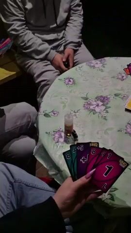 The video shows a close-up of a person's hand holding four UNO cards: a green 8, a purple +4, a pink 8, and a pink 1. The hand has long, pointed, pink acrylic nails. The cards are fanned out. In the background, a round table covered with a floral tablecloth is visible. On the table, there are more UNO cards, a green can of EFES beer, and a small clear bottle with a colorful lid. Two other people are partially visible sitting at the table, one wearing a gray hoodie and sweatpants, and the other wearing a gray sweatshirt and jeans. They appear to be playing UNO outdoors at night, with a dark background suggesting a yard or patio.