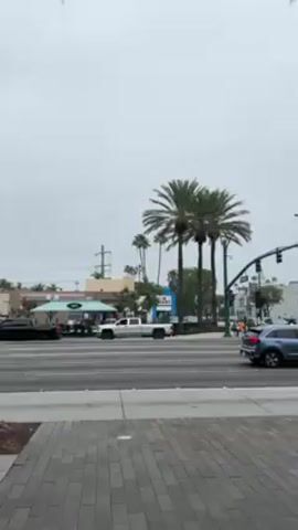 The camera stands stationary on a brick sidewalk at a busy street intersection on a cloudy day. Cars and buses proceed through the intersection. Tall palm trees line the street. There are several buildings in the background. A group of people stand on the corner waiting to cross the street.