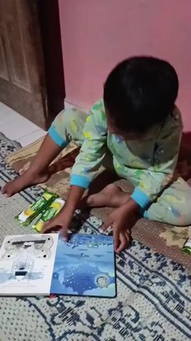 A young child wearing a light green pajama set with a cartoon pattern is sitting on a patterned rug. The child is looking down at an open book with colorful illustrations. To the child's left are two green and yellow snack packages. The child's right hand is touching the book. Another person's hands are visible in the lower right corner of the frame, also interacting with the book or the rug. The background shows a pink wall and a wooden door. The lighting appears to be artificial and dim, suggesting an indoor evening scene. The child shifts position and picks up a snack.