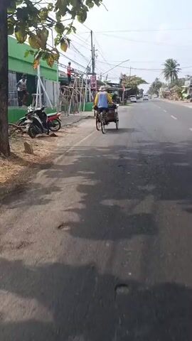 The video opens showing a side view of a person riding a three-wheeled bicycle with a covered passenger compartment, moving down a paved road. The rider wears a light-colored hat and a blue long-sleeved shirt with yellow sleeves. The bicycle has black tires with blue accents on the rims. To the left of the road, a building with a green facade is under construction or renovation. Scaffolding is erected along the building's side, and at least two individuals are visible on or near the scaffolding, possibly working. A red and black motorcycle is parked to the left. Further down the road, other vehicles and buildings are visible, suggesting a street in a town or village. The road surface appears to have some cracks and patches. The video continues with the perspective shifting to someone on a motorcycle, following the cyclist. The motorcycle rider's handlebars and dashboard are visible in the foreground. The view of the road ahead shows the cyclist in the distance, along with other traffic including cars and other motorcycles. The environment appears to be an outdoor, daytime scene with clear skies.