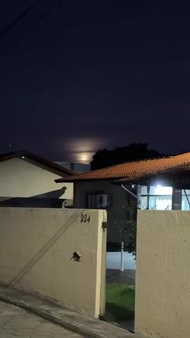 The video shows a dark, outdoor scene at night.  A bright moon is visible through a layer of clouds. The camera is focused on a cream-colored cement wall with a house behind it. The house has a brown tiled roof and an air conditioner unit is visible. The house number "224" is visible on the wall. A shadow of a bird is visible on the wall. The scene is lit by the moonlight and a light in the house.