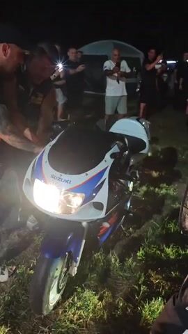 A group of people are gathered around a blue and white Suzuki motorcycle at night. The motorcycle's headlights are on. Some people are holding up their phones, seemingly to record or take pictures. The environment appears to be an outdoor event with grass and tents visible in the background. A person in the foreground on the right side of the frame is partially visible, wearing an orange and black jacket. Another person on the left side of the frame, wearing a dark t-shirt and hat, is reaching towards the motorcycle. The overall atmosphere suggests a gathering or event involving the motorcycle.