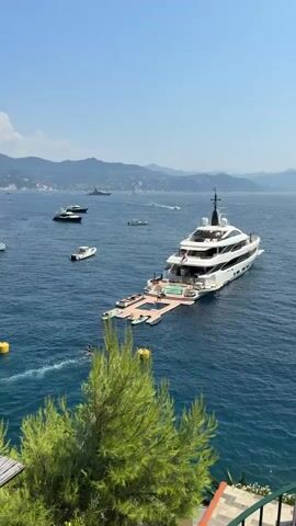 The video shows a large white yacht anchored in clear blue water, with mountains in the background. The yacht has a swimming pool and a large deck area extended from the stern, with several smaller boats and inflatable toys. A few people are visible on the deck, and some are swimming in the water near the yacht. Several other boats are also visible in the distance. The sky is clear and blue, indicating a sunny day. The foreground shows a lush green tree and part of a balcony with railings.