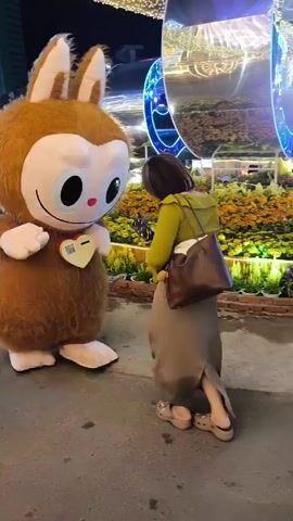 At night, a woman poses for a photo with a brown and white mascot in an outdoor public setting. The mascot, resembling a rabbit-like cartoon character with large eyes and a heart-shaped badge, stands on a paved surface. A woman in a green shirt and tan pants approaches the mascot and hugs it. She then steps back and takes a photo of the mascot with her phone. In the background are flower beds, illuminated decorations, buildings, and other people.