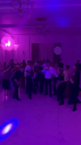 A group of people are dancing in a ballroom with a polished floor and white walls. They are illuminated by changing colored lights. There is a group of dancers in matching black and pink outfits in the foreground. A group of people are standing and watching in the background. The room is decorated with circular hanging lights.
