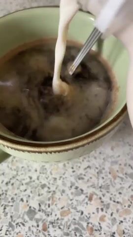 A person is pouring milk into a cup of coffee. The cup is light green with a darker green rim and handle and sits on a countertop patterned with beige, brown, and black specks. Steam rises from the coffee as the milk is poured from a small plastic bottle.