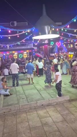 A nighttime outdoor scene shows a gathering of people, possibly at a festival or market, with colorful string lights draped overhead. A central area is paved with large, square stones, and a low stone wall borders the foreground. In the background, there are structures that appear to be stalls or shops adorned with more lights and some circular displays. Several individuals are engaged in various activities: some are standing and conversing, a few are seated on the low wall, and a group of people are dancing in the paved area. One person, wearing a white top and black and white striped pants, is facing away from the camera and appears to be leading a small child in red clothing. Other people are dancing in pairs or small groups. The atmosphere seems festive and social.
