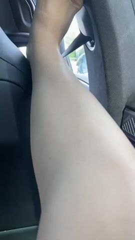 A person's arm is visible from the elbow down, resting near a car door. The interior of the car is dark with textured paneling. Outside the window, a highway can be seen with other cars passing by, lush green trees, and a cloudy sky. The perspective suggests the video is being filmed from inside a moving vehicle.