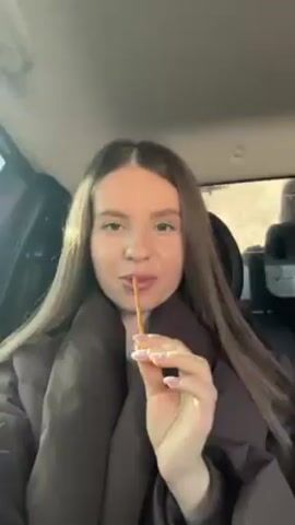 A woman is in the passenger seat of a car. She is wearing a brown puffer coat. She has long brown hair and is eating a pretzel stick. She looks at the camera and then looks out the window. The background is blurred, suggesting the car is moving. The lighting indicates it is daytime.