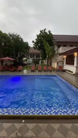 The video shows a rectangular swimming pool with blue water and a blue tiled edge. The pool is surrounded by lounge chairs. In the background, there is a building with a tiled roof and balconies, as well as trees and a covered parking area with a car. The sky is overcast. The pool is lit with blue underwater lights.