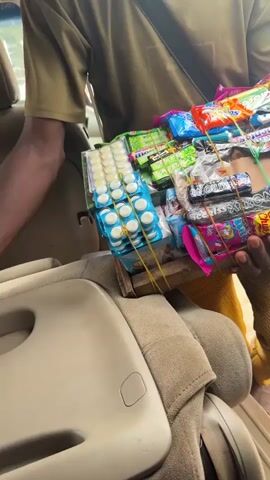 A person wearing a green shirt and yellow pants is holding a tray full of candy bars and other snacks. They are inside a vehicle, with the interior of the car visible in the background. The tray is made of wood and is secured with rubber bands. The person appears to be a vendor, possibly selling snacks to people in the car or on the street. The lighting is natural, suggesting it is daytime. The focus is on the person and the tray of snacks.