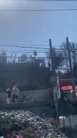 The video shows a scene of devastation, possibly after a fire or natural disaster. The terrain is hilly and covered with debris.  A group of three people are standing near a concrete structure, possibly a foundation. The vegetation appears burnt, with bare trees visible on the hillside. Chilean flags are erected amidst the rubble on the hill. The sky is clear and bright with the sun visible in the frame.  Electrical lines run across the top of the frame. A white car drives by on the street below.