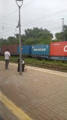 The video shows a train station platform on an overcast day. A freight train with numerous shipping containers is passing by. People are visible on the platform, some walking, some standing, and some interacting. One pair of individuals, a male and a female, are standing close together on the platform and appear to be in conversation. Another person, wearing a reflective vest, is walking on the platform. A person with a suitcase is also seen. The platform is paved with brick-like material, and a yellow line marks the edge. Trees and some buildings are visible in the background beyond the train tracks. The train cars are marked with company names such as "SINOKOR" and "CK LINE".