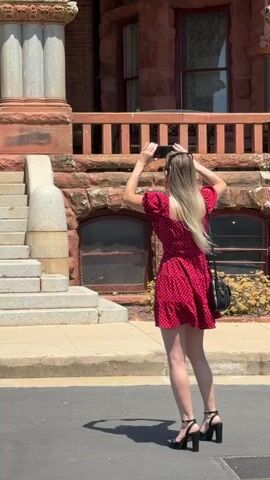 A young blonde woman in a red polka dot dress and black heels stands on a paved surface, holding a smartphone to take a photo of a large, red brick building. She is positioned to the right of a set of stone stairs leading up to an arched entrance of the building. A couple, a man and a woman, are visible on the stairs, facing the camera. The building has multiple windows and decorative stonework. The sky is clear and blue, indicating a sunny day. The perspective of the shot changes as the video progresses, zooming out to reveal more of the building's facade and the surrounding area.