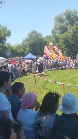 A person dressed in a traditional outfit, possibly a man, walks across a grassy field carrying two buckets, one in each hand, suspended from a yoke across their shoulders. The person is walking towards the right of the frame, and the buckets appear to be full of liquid, as evidenced by the splashing. They are participating in a competition or performance, as indicated by the crowd seated on the grass watching them. The background features a large, inflatable, multi-colored slide, tents, trees, and a clear blue sky, suggesting an outdoor festival or event. Colorful triangular flags are strung across the field, further indicating a celebratory atmosphere. The crowd is diverse in age and gender, and many are looking intently at the person carrying the buckets. The focus is on the person's movement and the action of carrying the buckets.