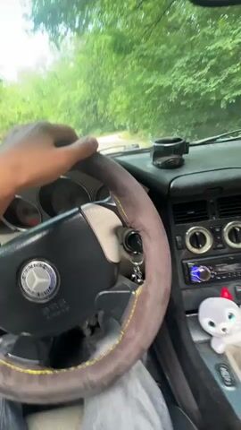 The video shows a person driving a car, originating from Cherganovo, Bulgaria.

The initial shot is from inside the car, showing a driver's hand on a steering wheel. The steering wheel has a grey, possibly suede, cover with yellow stitching and features a Mercedes-Benz logo. Parts of the dashboard are visible, including a small white toy with red accents on the passenger side. Through the windshield, green trees and foliage are seen, indicating an outdoor, possibly rural or suburban setting.

The perspective then shifts to an outward view from the car, showing the vehicle entering or driving through a concrete underpass or tunnel. The rear deck of the car is visible, appearing to be a teal or turquoise color, fitted with a prominent black spoiler. The concrete walls of the underpass have some graffiti. As the car moves through the underpass, a loud engine or exhaust sound is heard, presumably from the car accelerating or revving its engine, creating an echo effect. Bright light is visible at the end of the tunnel, indicating it leads back to the open road. The scene takes place during the daytime.