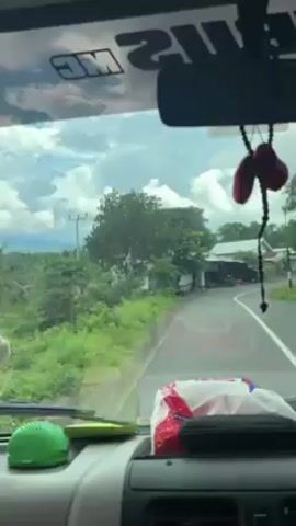 A person is driving down a paved road in a vehicle. The road is lined with trees and vegetation on either side. There are some houses and a power line visible in the background. The sky is cloudy. There is a small rosary hanging from the rearview mirror. The driver is speaking in Indonesian.