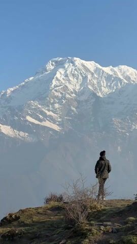 A person stands facing a large, snow-capped mountain under a clear blue sky. The person is wearing a dark jacket, pants, and a beanie. They are standing on a grassy hill with some small shrubs in the foreground. The mountain dominates the background, its peaks covered in snow and its slopes partly visible through a misty haze. The person is viewed from the back, and they are stationary, seemingly admiring the mountain. There are voices heard in the background.
