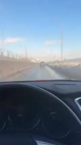 The video, taken from inside a car, shows a view through the front windshield while driving on a highway. A car is visible in the distance, ahead on the highway. There is a bright blue sky with a few clouds. Light posts are visible on the side of the road. There is a concrete barrier on the left side of the road. The dashboard and steering wheel of the car are visible in the foreground. The road appears wet.
