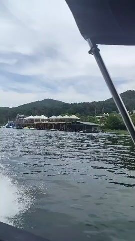 The video shows a boat moving on the water. The camera is positioned on the boat, looking forward and to the right. The water is choppy and white foam is visible from the boat's wake. In the distance, there is a large structure on the water with a white roof. Behind the structure, there are hills covered in trees. The sky is cloudy.