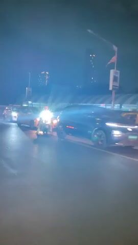 The video shows a nighttime scene on a busy road with several vehicles. In the foreground, a scooter is moving forward, carrying two people. The rider wears a helmet, and the passenger is dressed in a patterned outfit with a light-colored scarf. In the middle ground, there are several cars, some of which have their headlights on, illuminating the road. One car appears to be stopped or moving slowly in traffic. The background reveals city lights and some structures, including what looks like a covered walkway or bridge. The overall impression is of urban traffic at night.