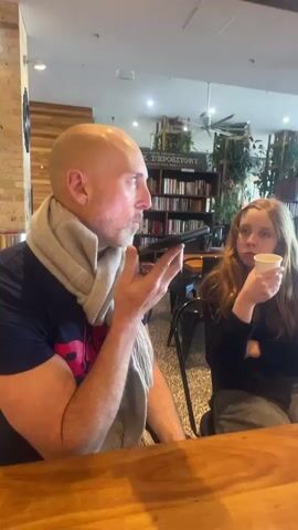 A bald man wearing a grey scarf and a dark t-shirt with a red design is holding a black smartphone up to his ear and speaking. He is seated at a wooden table. To his right, a young woman with long red hair is holding a white paper cup to her lips. She is wearing a black top. Behind them, shelves filled with books are visible, along with some plants and ceiling fans. The setting appears to be a cafe or library.