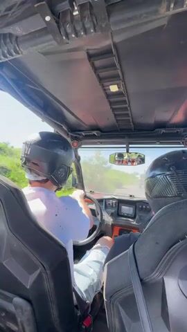 Two people wearing helmets are riding in a side-by-side vehicle. The person in the driver's seat is wearing a white t-shirt and ripped jeans. The person in the passenger seat is wearing a black shirt. They are driving on a dirt road with grassy hills on either side. The sky is blue and there are some trees in the distance. The vehicle is moving forward.